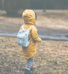 A preschool aged child walks in the grass with a yellow jacket on and a blue backpack