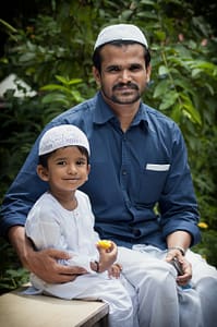 A father and young child smile at the camera while eating fruit in a garden