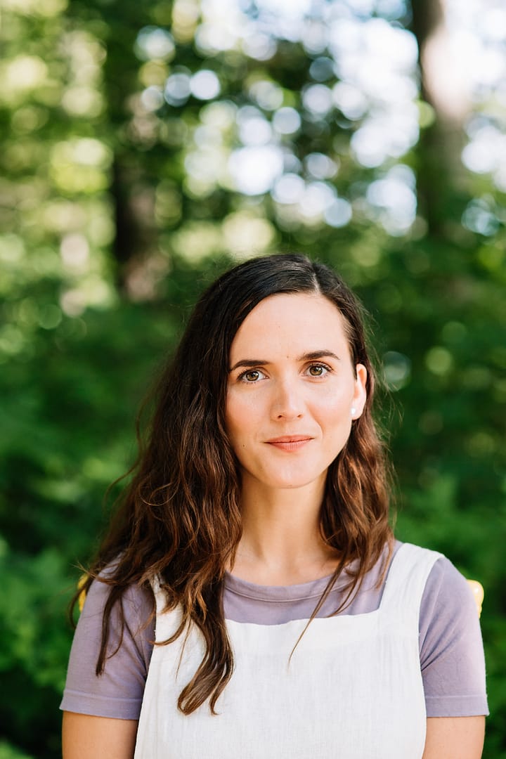 Robin, Darling+ Founder and experienced child care provider, smiles at the camera, wearing white overalls over a purple t-shirt. There are trees in the background.