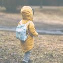 A preschool aged child walks in the grass with a yellow jacket on and a blue backpack